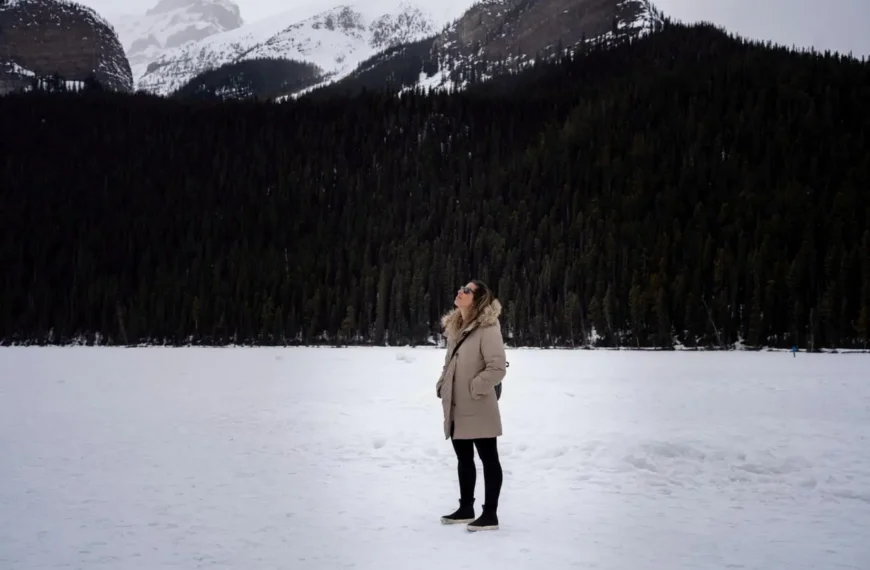 Mulher com roupas de frio em um parque cheio de neve e ao fundo uma floresta