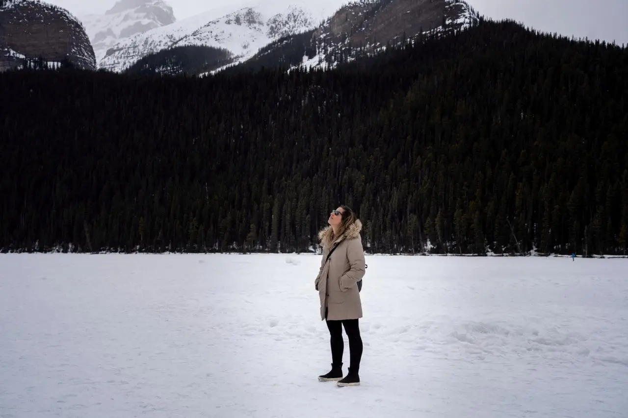 Mulher com roupas de frio em um parque cheio de neve e ao fundo uma floresta