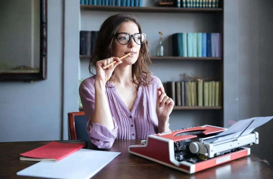 Retrato de uma escritora madura, pensativa, usando óculos e sentada em frente a uma máquina de escrever vermelha